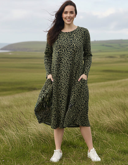 A woman wearing a long-sleeve olive green midi dress with a black leopard print pattern and white sneakers, standing in a grassy field with rolling hills under a cloudy sky.