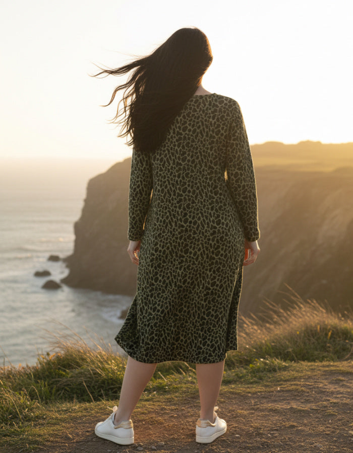 A woman stands on a cliffside path at sunset wearing a long-sleeve olive green and black leopard-print midi dress with white sneakers, with the ocean and rocky coastline in the background.