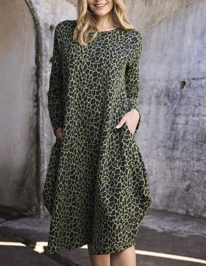 A woman wearing a long-sleeve olive green dress with a black leopard print pattern, standing in front of a textured concrete wall.
