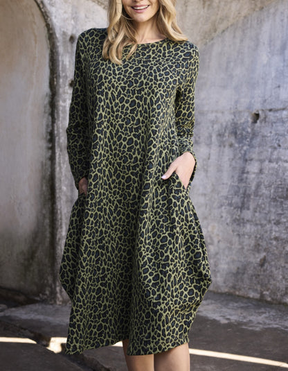A woman wearing a long-sleeve olive green dress with a black leopard print pattern, standing in front of a textured concrete wall.