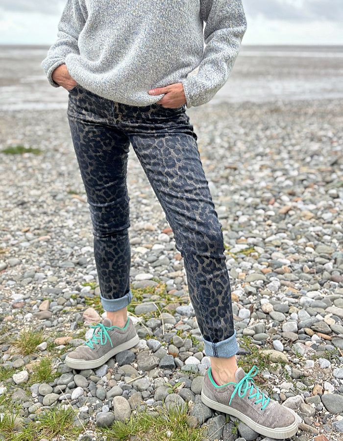 Person wearing leopard print pants and gray sneakers on a pebbly beach.