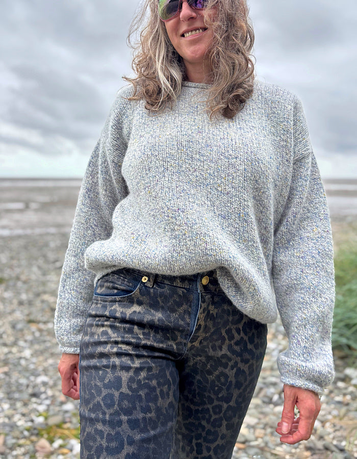 Woman wearing a gray sweater and patterned animal print jeans standing on a beach.