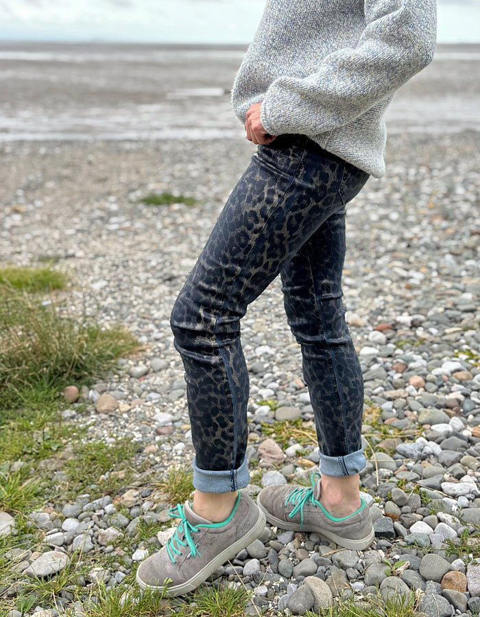 Person wearing leopard print jeans and gray sneakers on a pebbly beach.