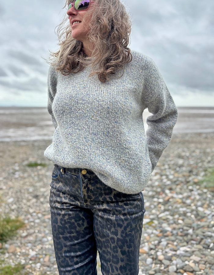 Woman wearing a light gray sweater and leopard print pants standing on a pebbly beach.
