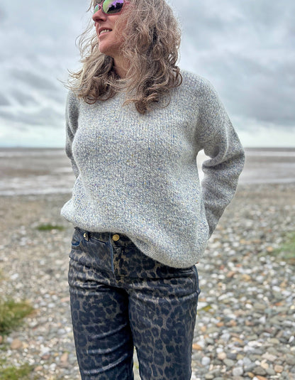 Woman wearing a light gray sweater and leopard print pants standing on a pebbly beach.