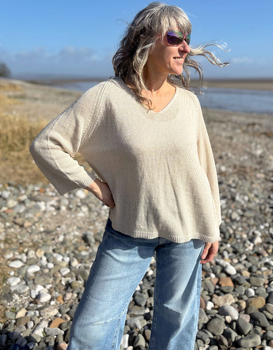 Woman standing on a pebbly beach wearing sunglasses and a beige sweater.
