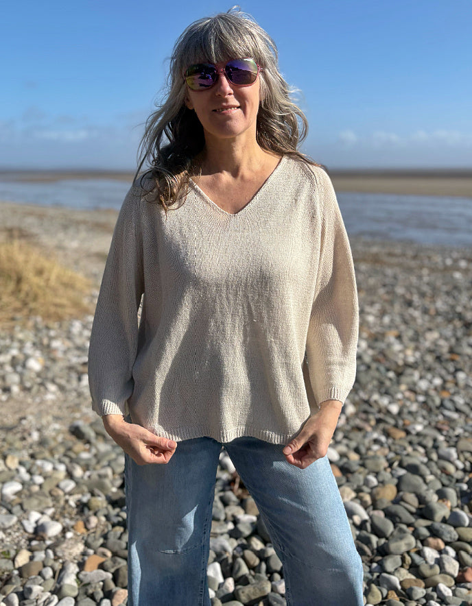 Woman standing on a pebbly beach wearing sunglasses and a beige linen knit sweater.