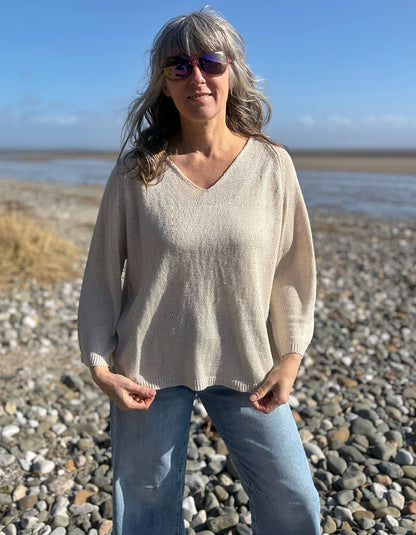 Woman standing on a pebbly beach wearing sunglasses and a beige linen knit sweater.