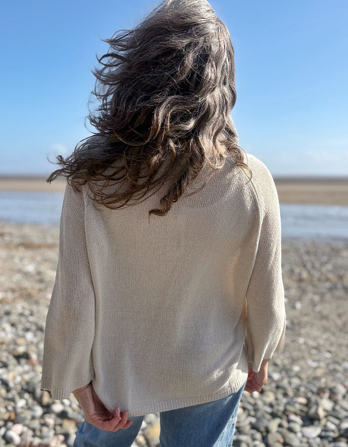 Woman standing on a pebbly beach wearing sunglasses and a cream linen knit sweater.