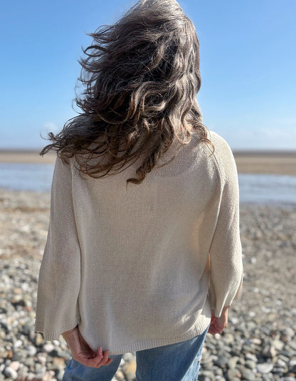 Woman standing on a pebbly beach wearing sunglasses and a cream linen knit sweater.