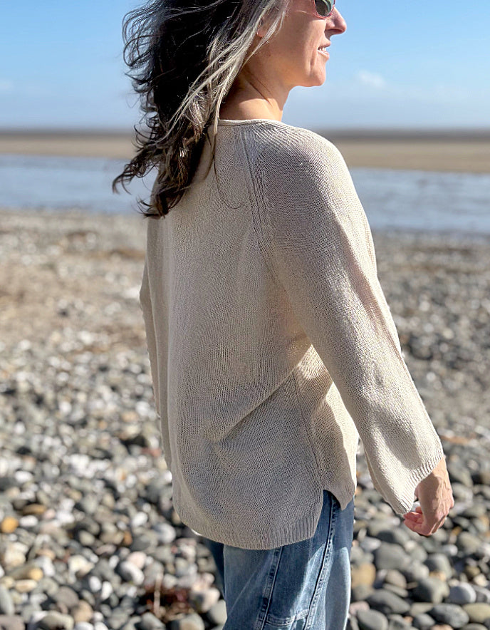 Woman standing on a pebbly beach wearing sunglasses and a cream linen knit sweater.