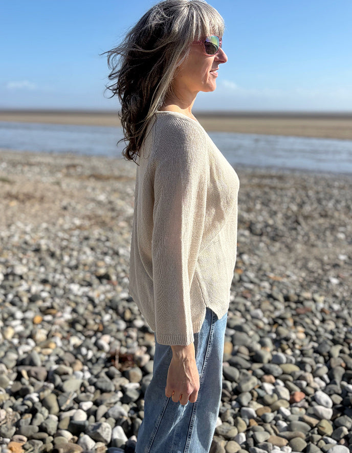 Woman standing on a pebbly beach wearing sunglasses and a cream linen knit sweater.