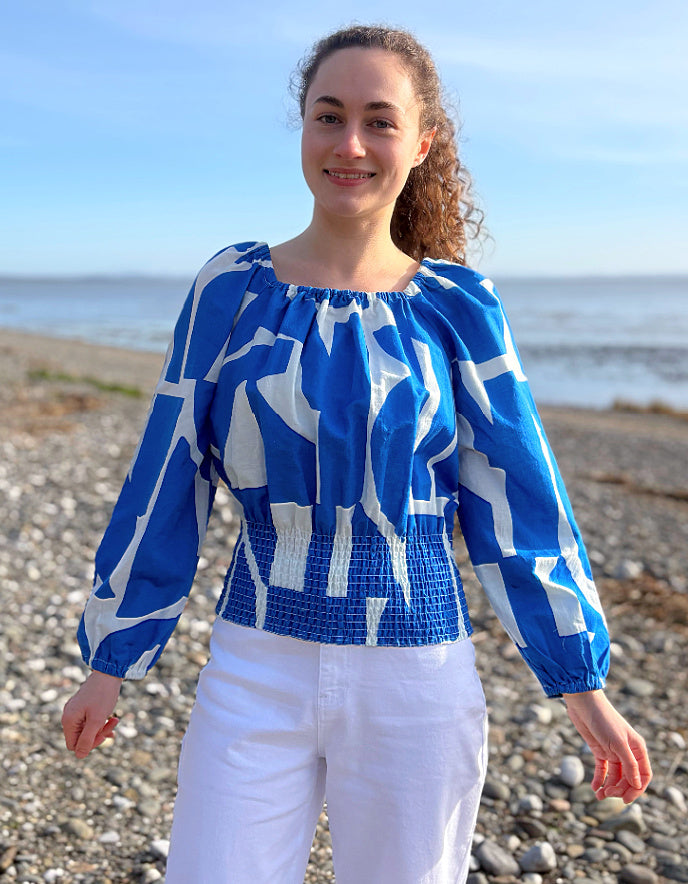 Woman wearing a blue and white patterned blouse standing on a pebbly beach.