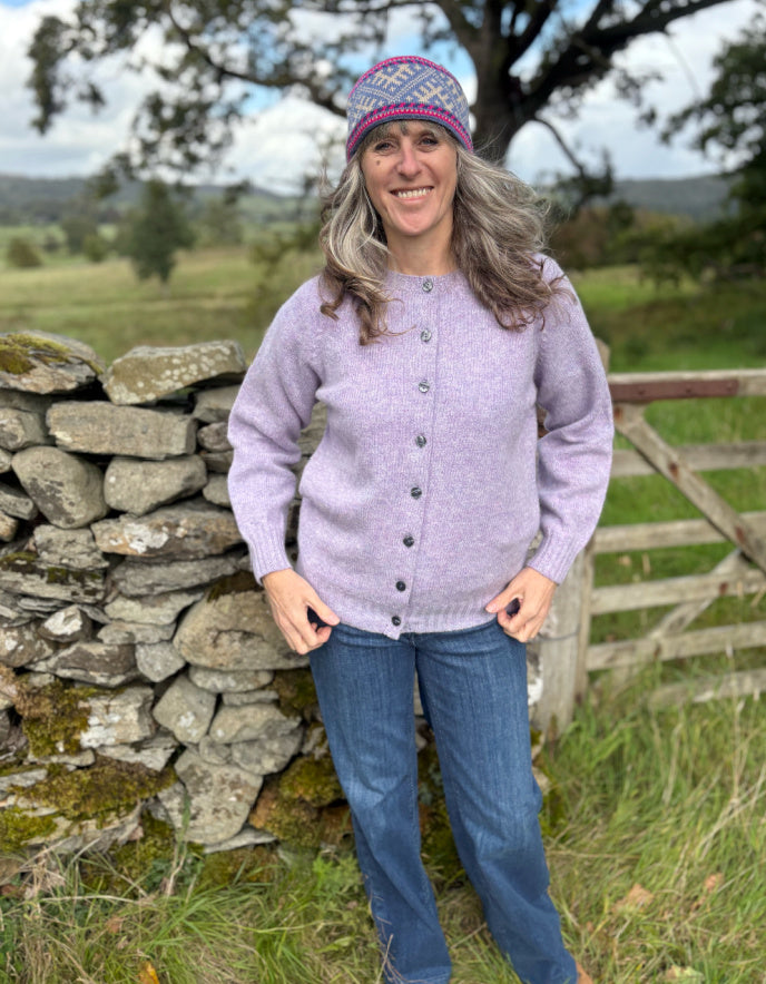 Woman wearing a purple sweater and knit hat standing in a field with a stone wall and wooden fence.