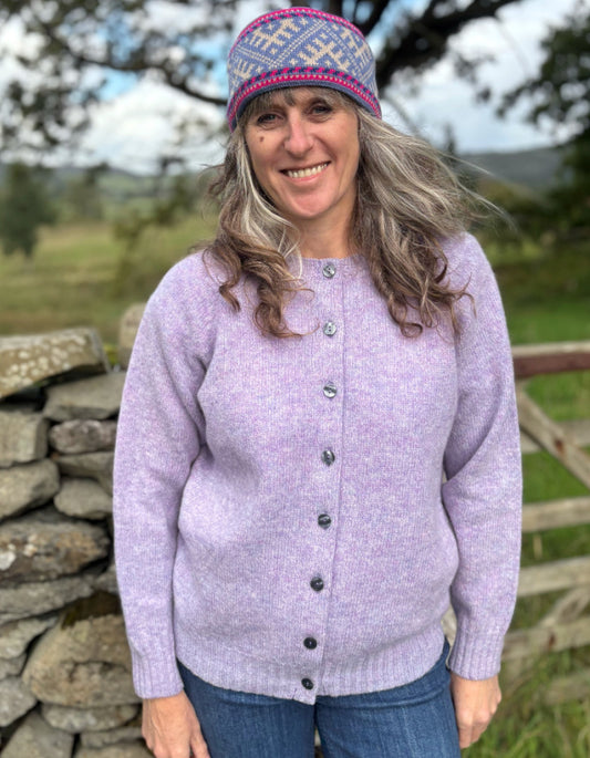 Woman wearing a purple cardigan and patterned hat outdoors near a stone wall.