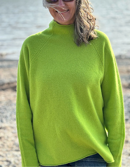 A woman wearing a bright lime green mock neck sweater, photographed outdoors with a blurred shoreline background.