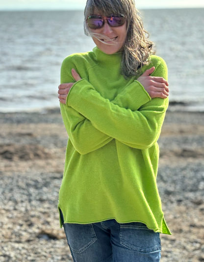 A woman wearing a bright lime green turtleneck sweater and blue jeans stands on a pebble beach with the water in the background, holding her arms close to her body.