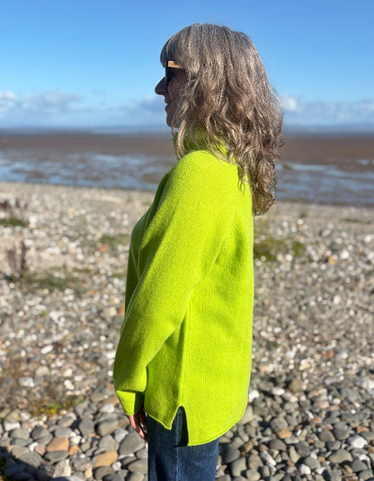 A woman with shoulder-length wavy gray-blonde hair stands in profile on a pebble beach, wearing a bright neon yellow-green knit sweater and dark blue jeans against a background of blue sky and water.