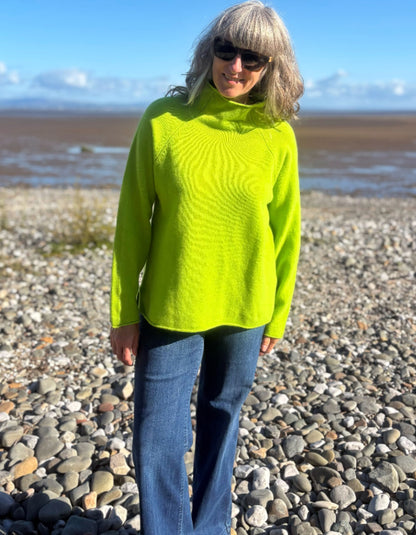 A woman wearing a bright neon yellow-green ribbed turtleneck sweater and blue jeans stands on a pebble beach with water and mountains in the background.