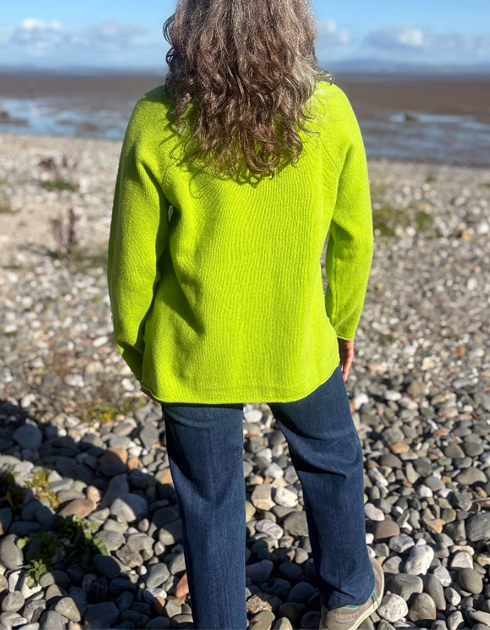 A person standing on a pebble beach, viewed from behind, wearing a bright neon yellow-green ribbed knit sweater and dark blue jeans.