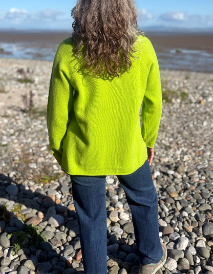 A person standing on a pebble beach, viewed from behind, wearing a bright neon yellow-green ribbed knit sweater and dark blue jeans.