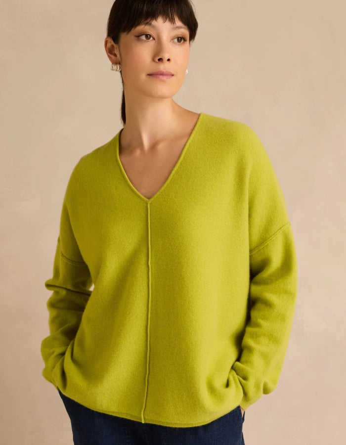 A woman wearing a bright lime green V-neck knit sweater with dropped shoulders, paired with dark blue jeans, standing against a neutral beige background.