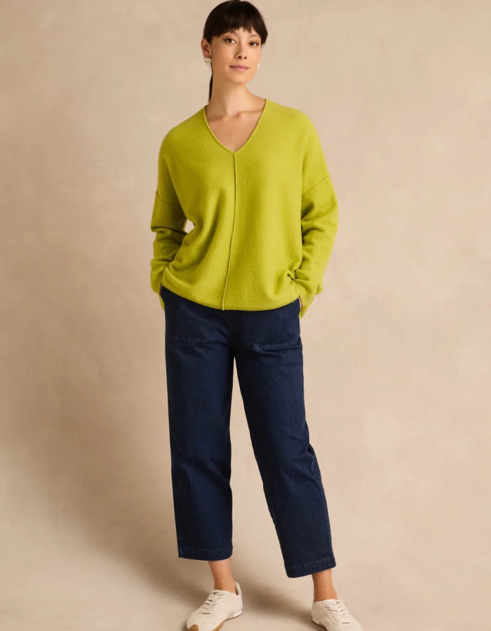A woman modeling a bright lime green knit cardigan sweater with a split V-neckline, paired with dark blue cropped pants and white sneakers, set against a warm beige studio backdrop.