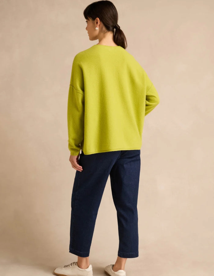A woman shown from the back wearing a bright lime green crewneck sweater and dark navy cropped pants, standing against a neutral beige studio backdrop.