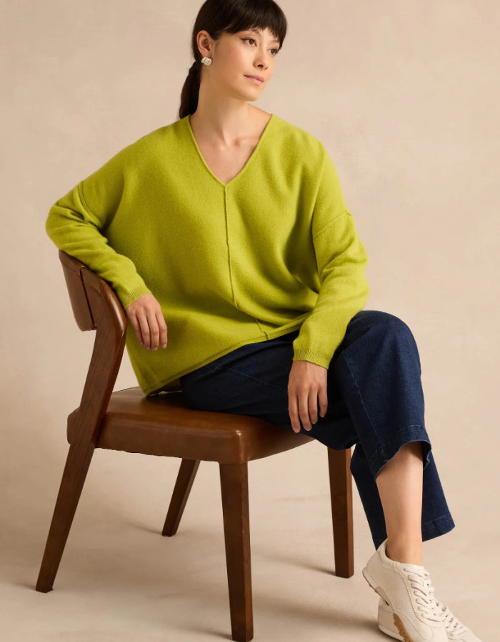 A woman sits on a wooden chair wearing an oversized lime green V-neck knit sweater, dark blue cropped pants, and white sneakers against a warm beige studio background.