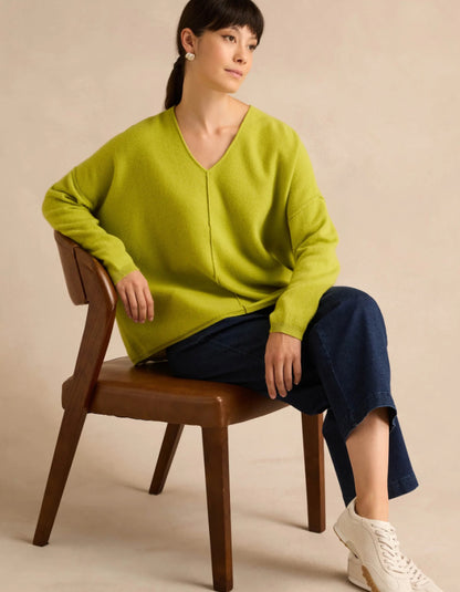 A woman sits on a wooden chair wearing an oversized lime green V-neck knit sweater, dark blue cropped pants, and white sneakers against a warm beige studio background.