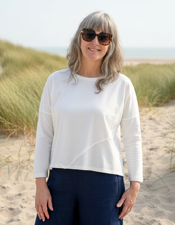 Woman wearing a white top and sunglasses on a sandy beach with grasses.
