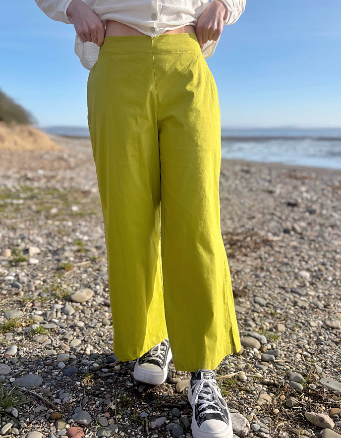 Person wearing bright lime wide leg pants and a white shirt on a pebbly beach.