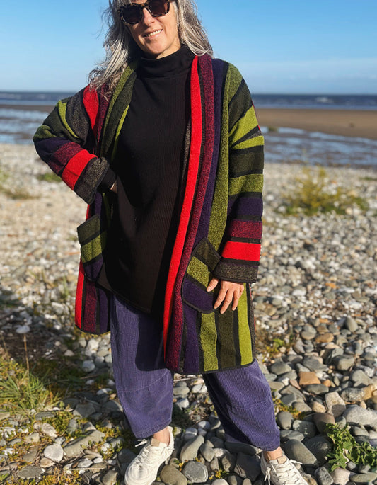 A woman standing on a pebble beach wearing a long striped cardigan with olive green, red, purple, and black stripes over a black turtleneck sweater, purple pants, and white sneakers.
