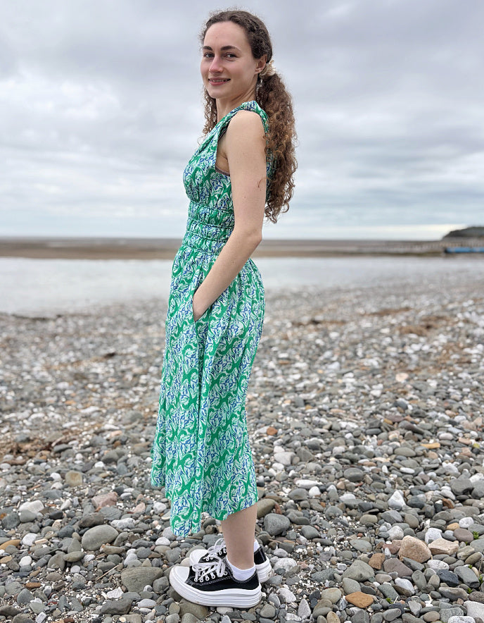 Woman in a green floral dress standing on a pebbly beach with a cloudy sky.