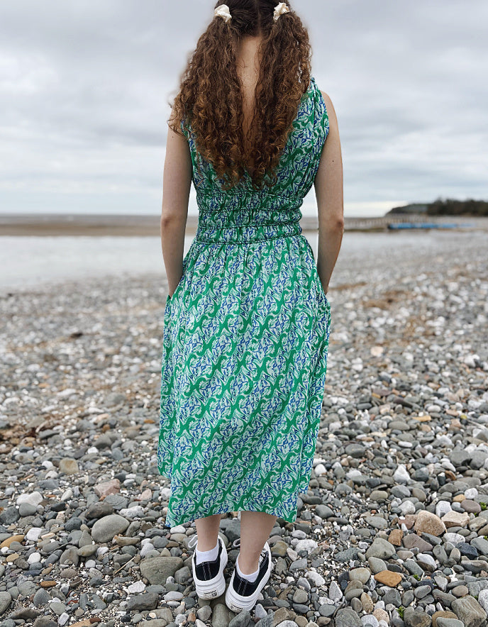 Person wearing a green patterned dress standing on a pebbly beach.