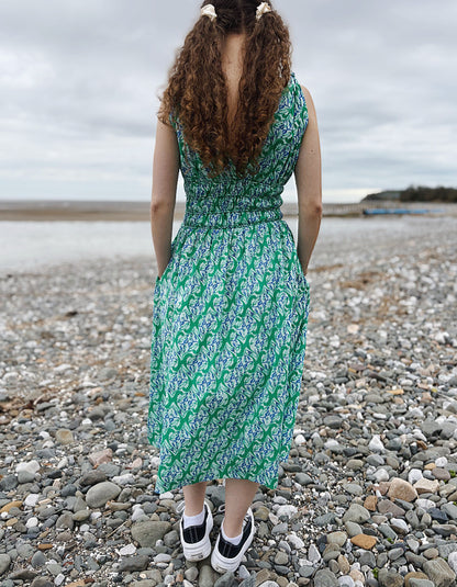 Person wearing a green patterned dress standing on a pebbly beach.