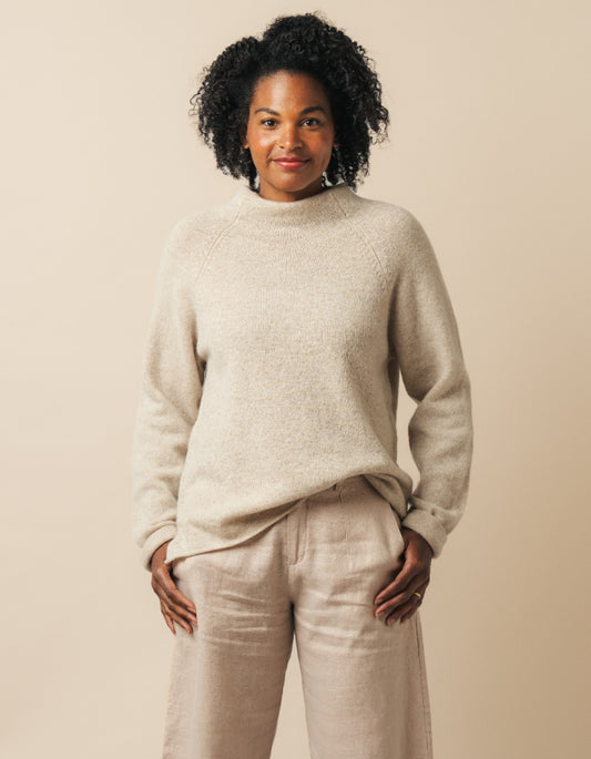 A woman with curly black hair wearing a light beige knit mock-neck sweater and matching beige trousers, standing against a solid beige background.