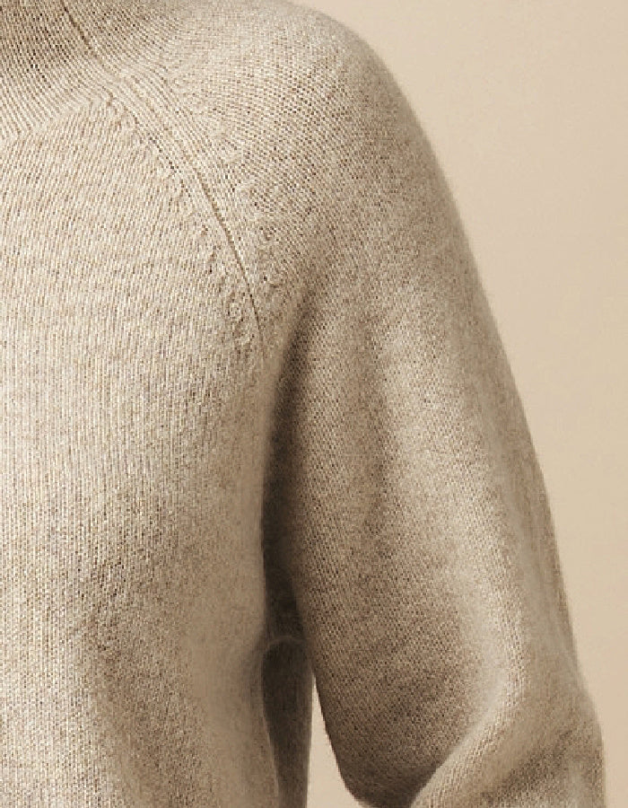 Close-up of a beige knit sweater showing the shoulder, sleeve, and ribbed texture against a matching neutral background.