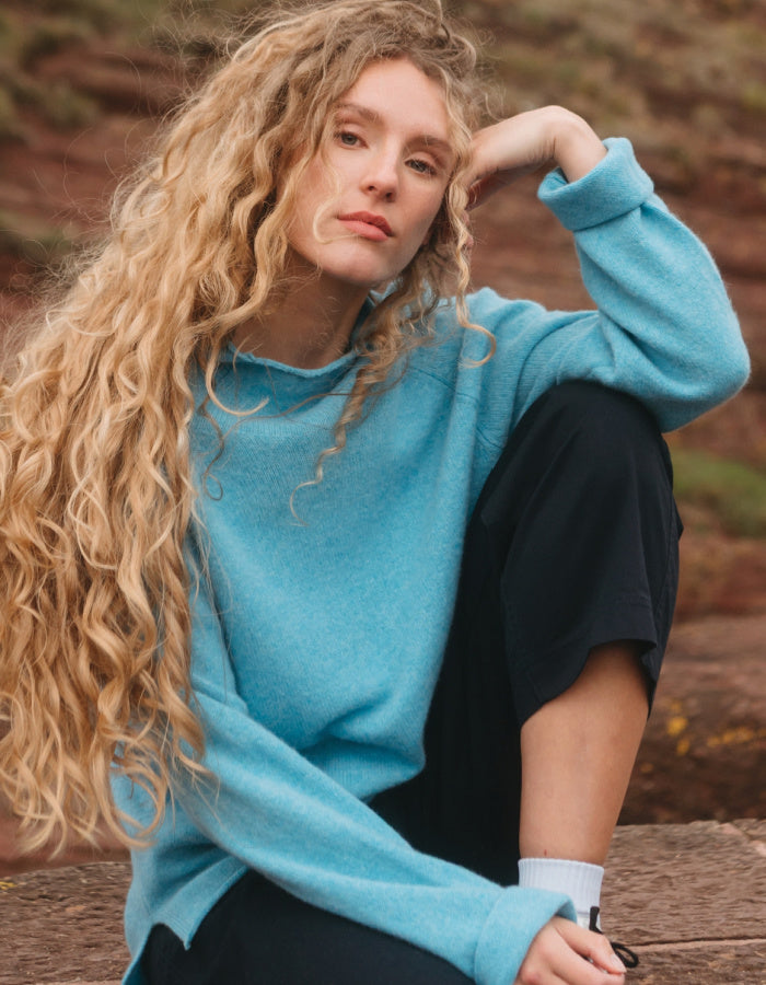 A woman with long, curly blonde hair wearing a bright turquoise blue knit sweater and black sweatpants, sitting outdoors on a wooden surface with a blurred natural background.