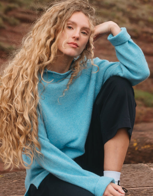 A woman with long, curly blonde hair wearing a bright turquoise blue knit sweater and black sweatpants, sitting outdoors on a wooden surface with a blurred natural background.