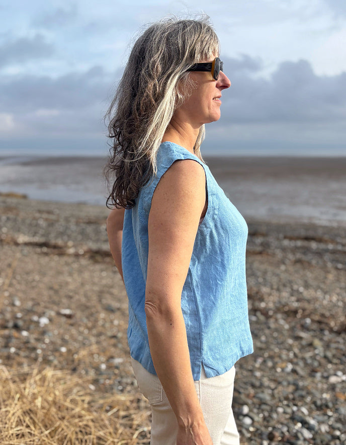 Woman standing on a beach wearing a blue sleeveless top and sunglasses, looking out towards the water.