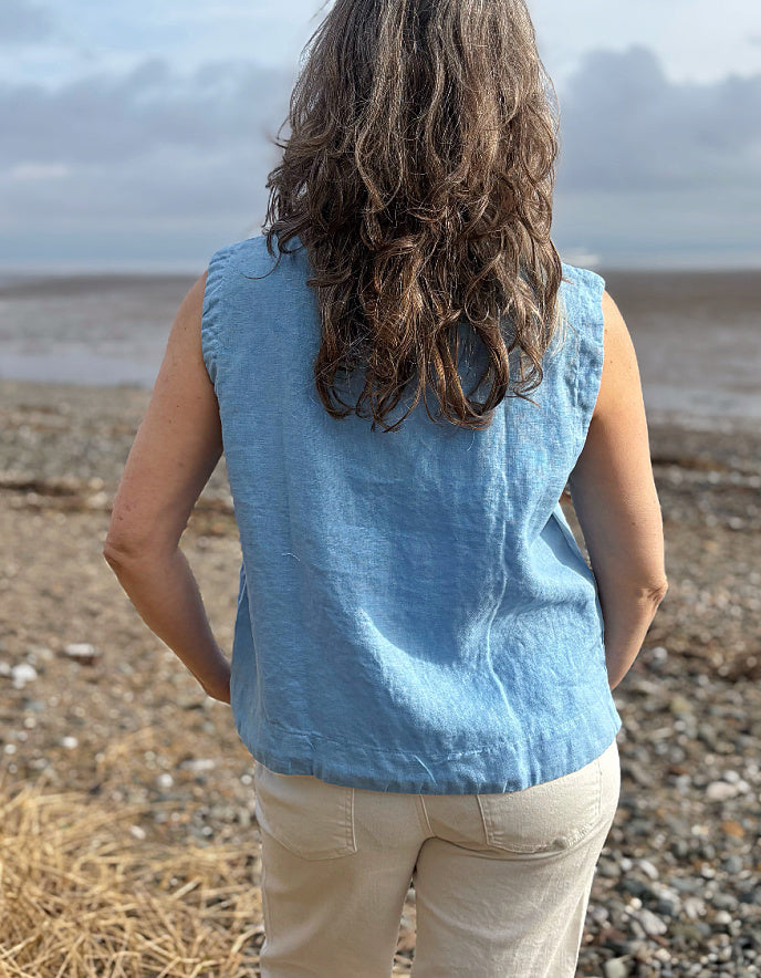 Person wearing a blue sleeveless shirt standing on a pebbly beach with a cloudy sky.