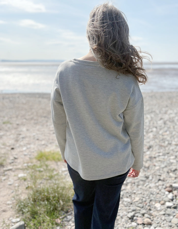 Person wearing a light gray sweater standing on a pebbly beach with ocean view