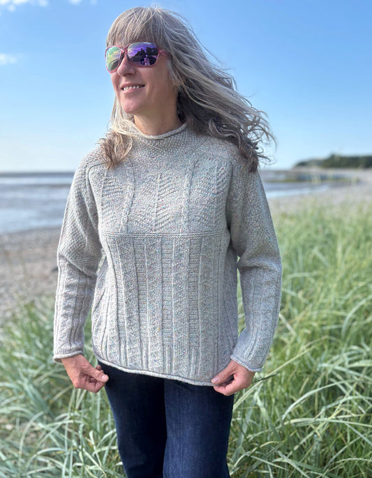 Person wearing a donegal tweed wool grey sweater with wide leg jeans and sunglasses on a beach