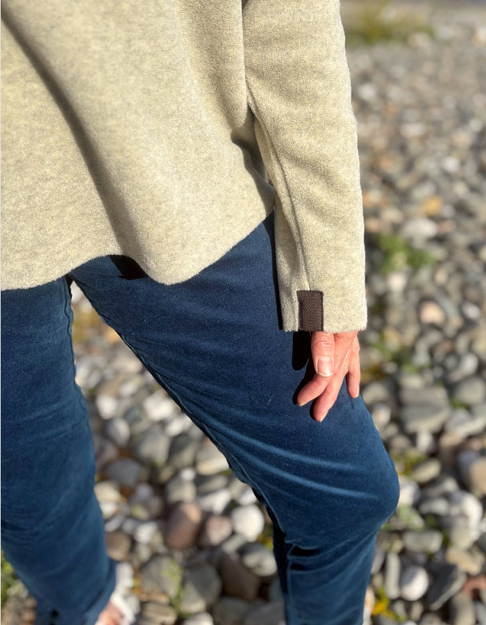 A close-up lifestyle shot of a person wearing a light beige knit sweater and dark teal velvet jeans, standing outdoors on a pebble-strewn shore. The person's hand is resting on the hem of the sweater, which features a small dark brown leather tab near the cuff.