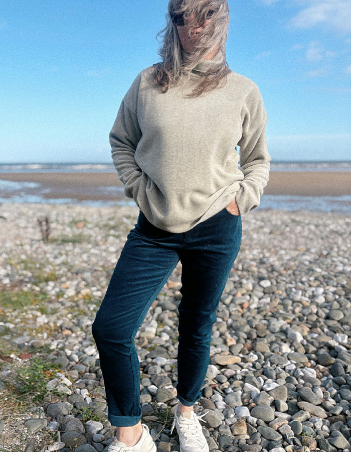 A woman standing on a rocky beach wearing a light beige knit sweater, dark teal velvet pants, and white sneakers, with the ocean and blue sky in the background.