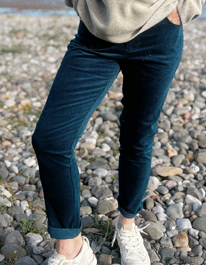 A woman standing on a rocky beach wearing a light beige knit sweater, dark teal velvet pants, and white sneakers, with the ocean and blue sky in the background.