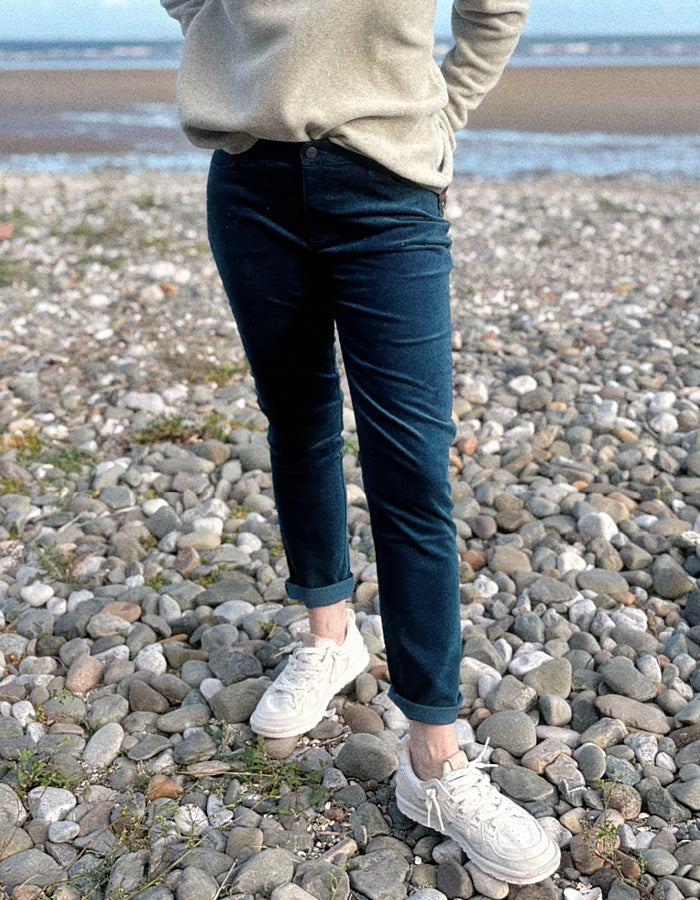 A woman standing on a rocky beach wearing a light beige knit sweater, dark teal velvet pants, and white sneakers, with the ocean and blue sky in the background.