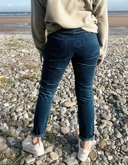 A person standing on a pebble beach, viewed from behind, wearing dark blue slim-fit teal velvet jeans, a beige knit sweater, and white slip-on sneakers.
