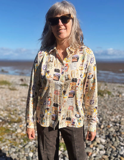 A woman with gray hair and sunglasses stands on a rocky beach wearing a beige button-up silky shirt covered in colourful illustrated print and dark brown cord wide leg pants, with water and blue sky in the background.